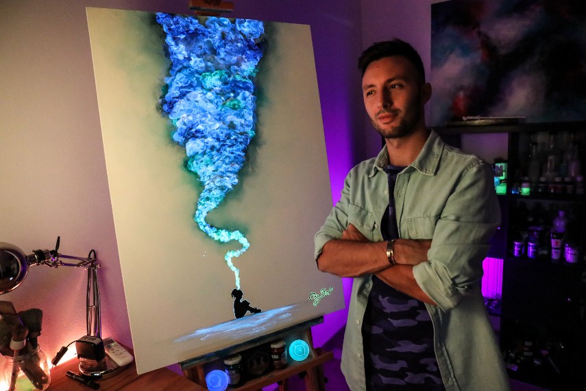 Photo of the artist in front of his painting, presenting the art that can glow in dark due to paint availabe to shop on youtube 